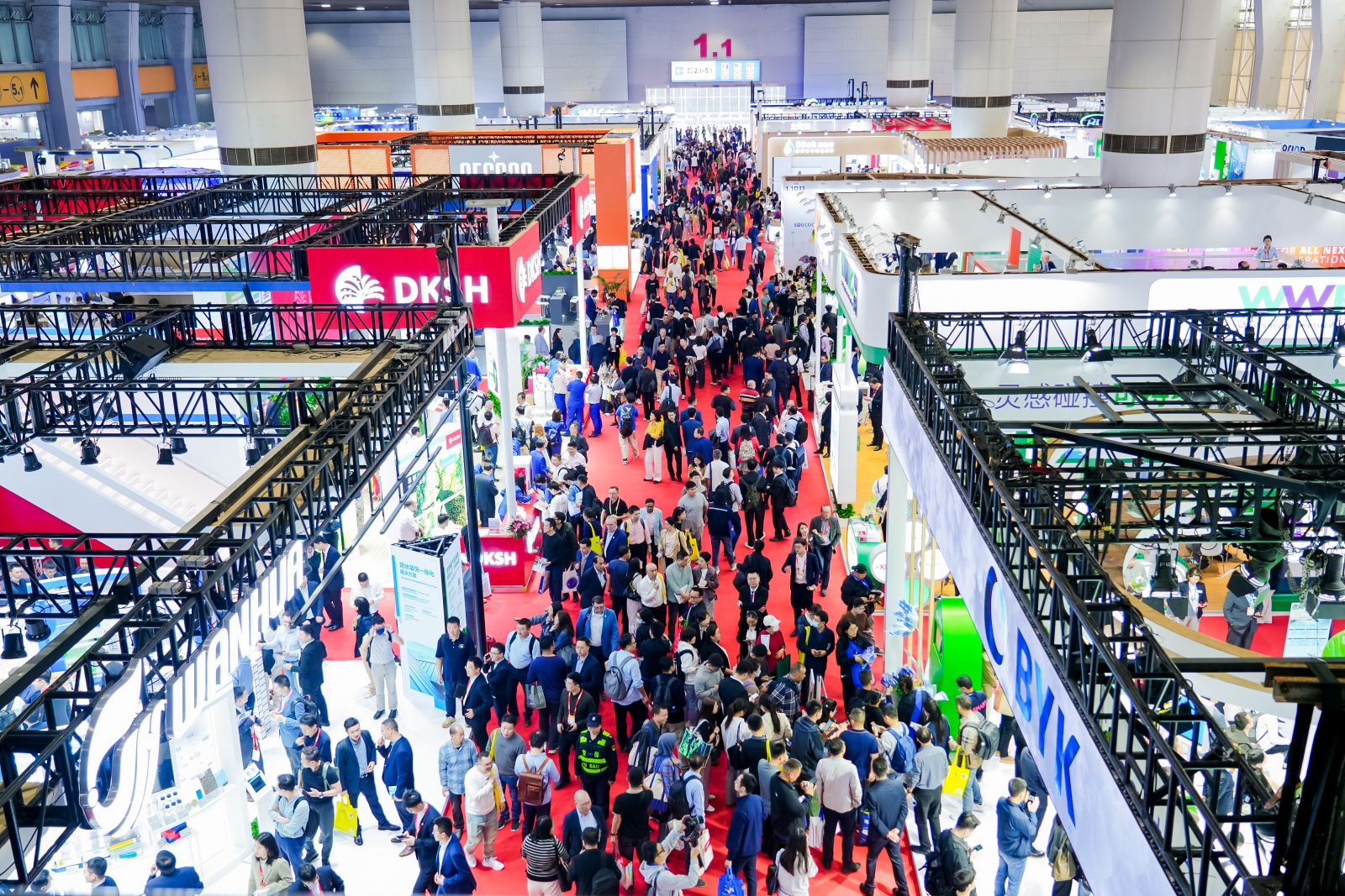Busy international trade exhibition hall filled with visitors, exhibition booths, red-carpet aisles, and large overhead lighting and display structures.