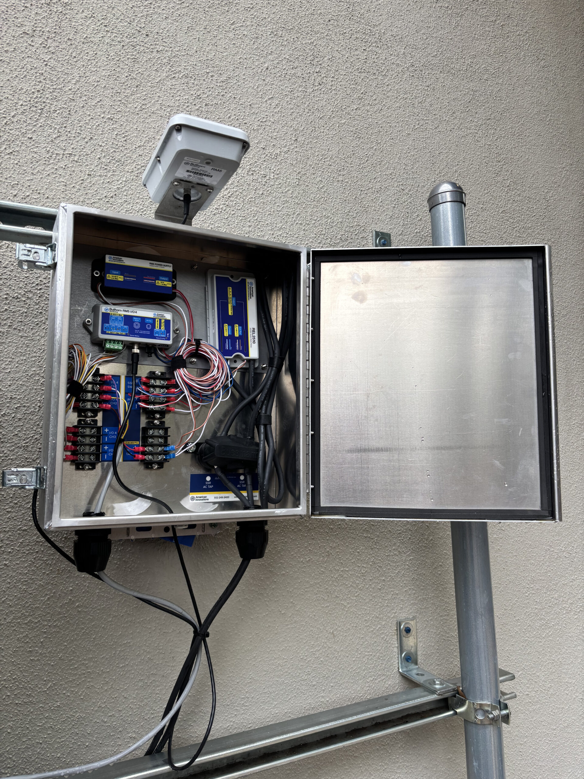 Industrial electrical control panel installed outdoors, with wiring, monitoring equipment, sensors, and electrical components visible inside an open metal enclosure.