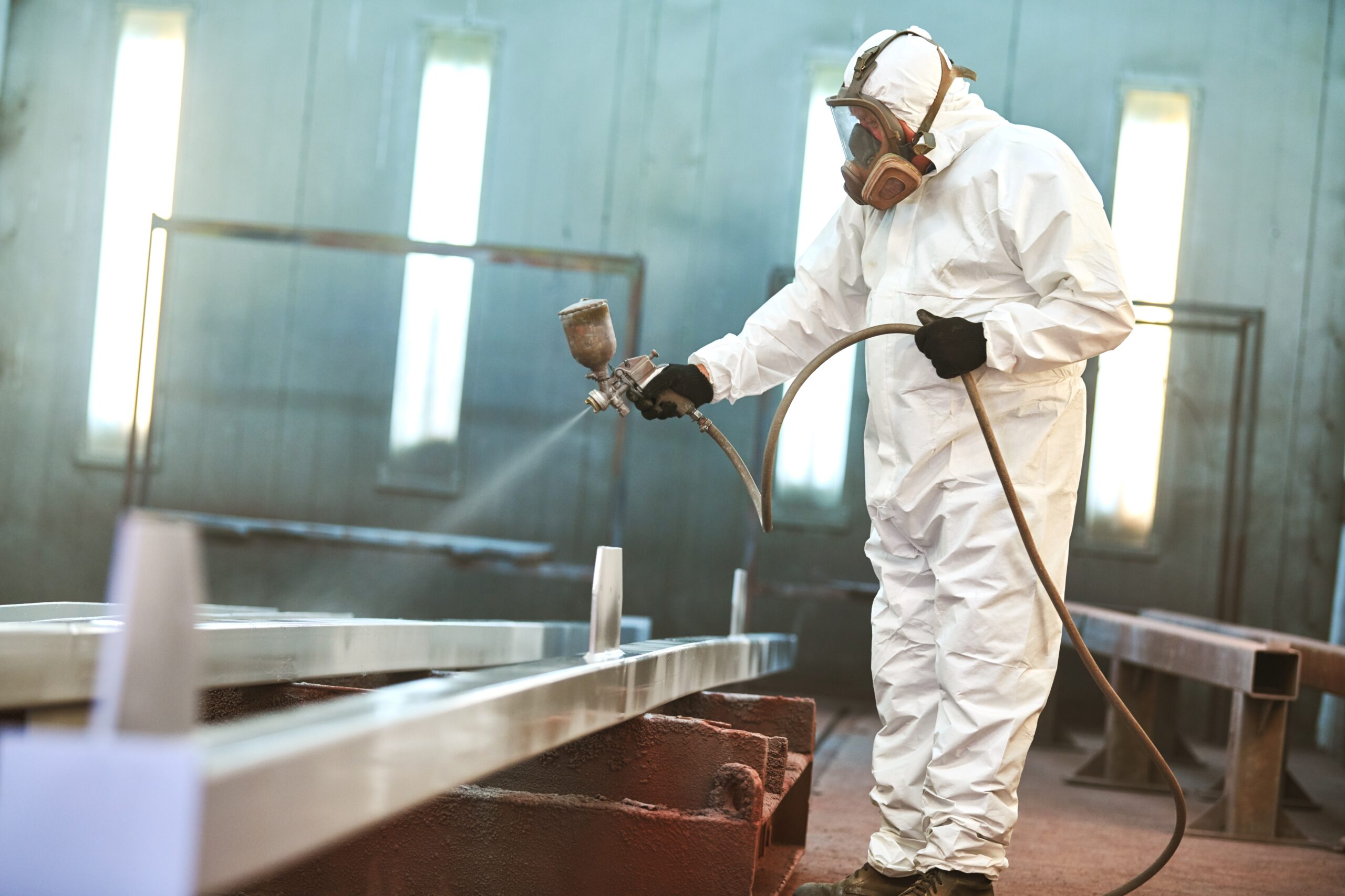 Professional industrial painter in protective coveralls and respirator spraying a coating onto a large metal structure inside a paint booth.