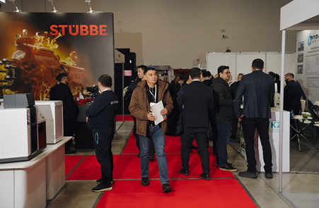 Visitors walking through a trade exhibition with company booths and industrial equipment displays along a red-carpeted aisle.