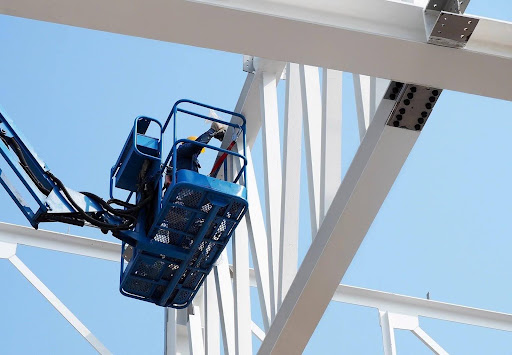 Worker in an elevated platform inspecting or working on white structural steel beams against a clear blue sky.