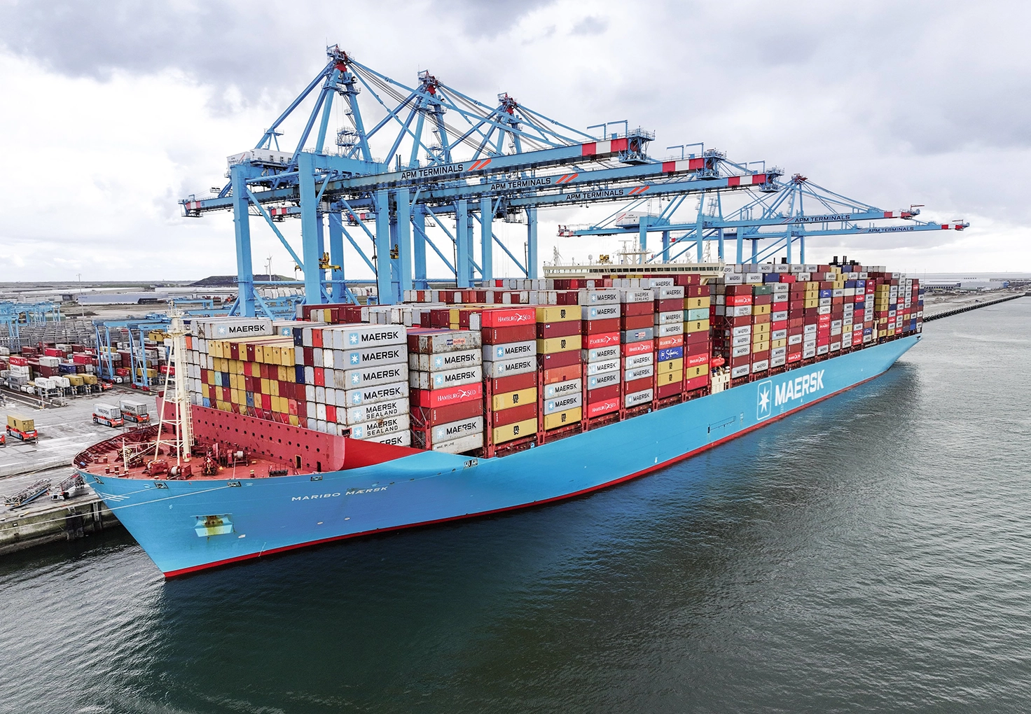 Container ship loaded with colorful cargo containers at a busy port terminal, with large container cranes in the background.